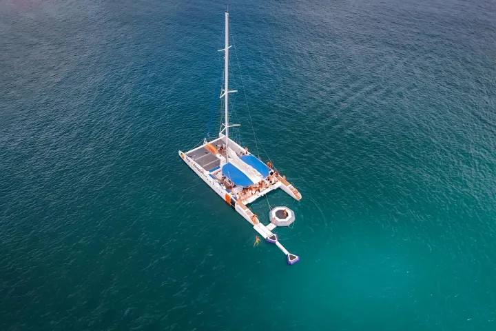 Aerial view of a catamaran with a blue canopy sailing on calm, clear blue ocean waters.